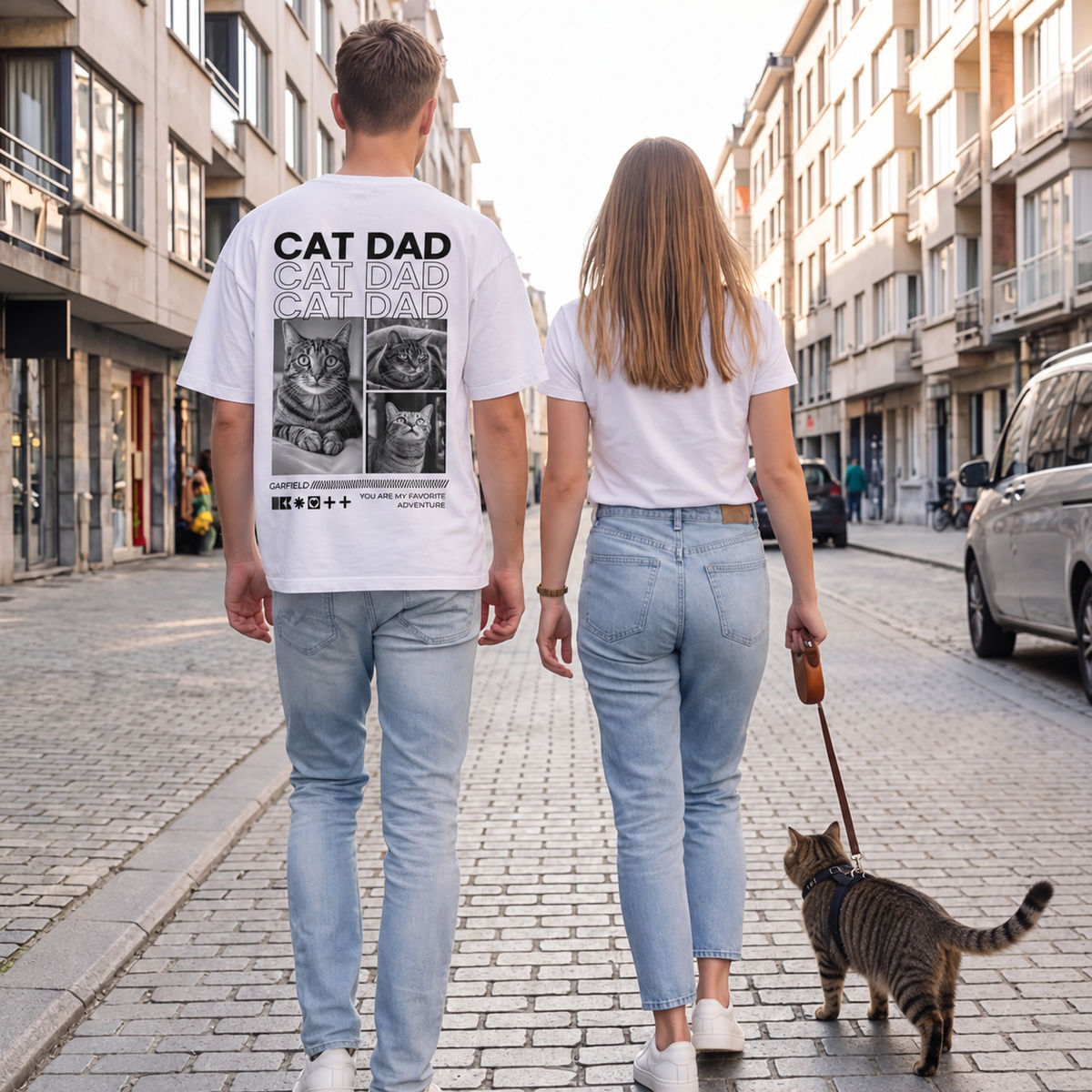 T-shirt bianca con stampa di gatti sul retro, due persone e un gatto a passeggio in una strada.
