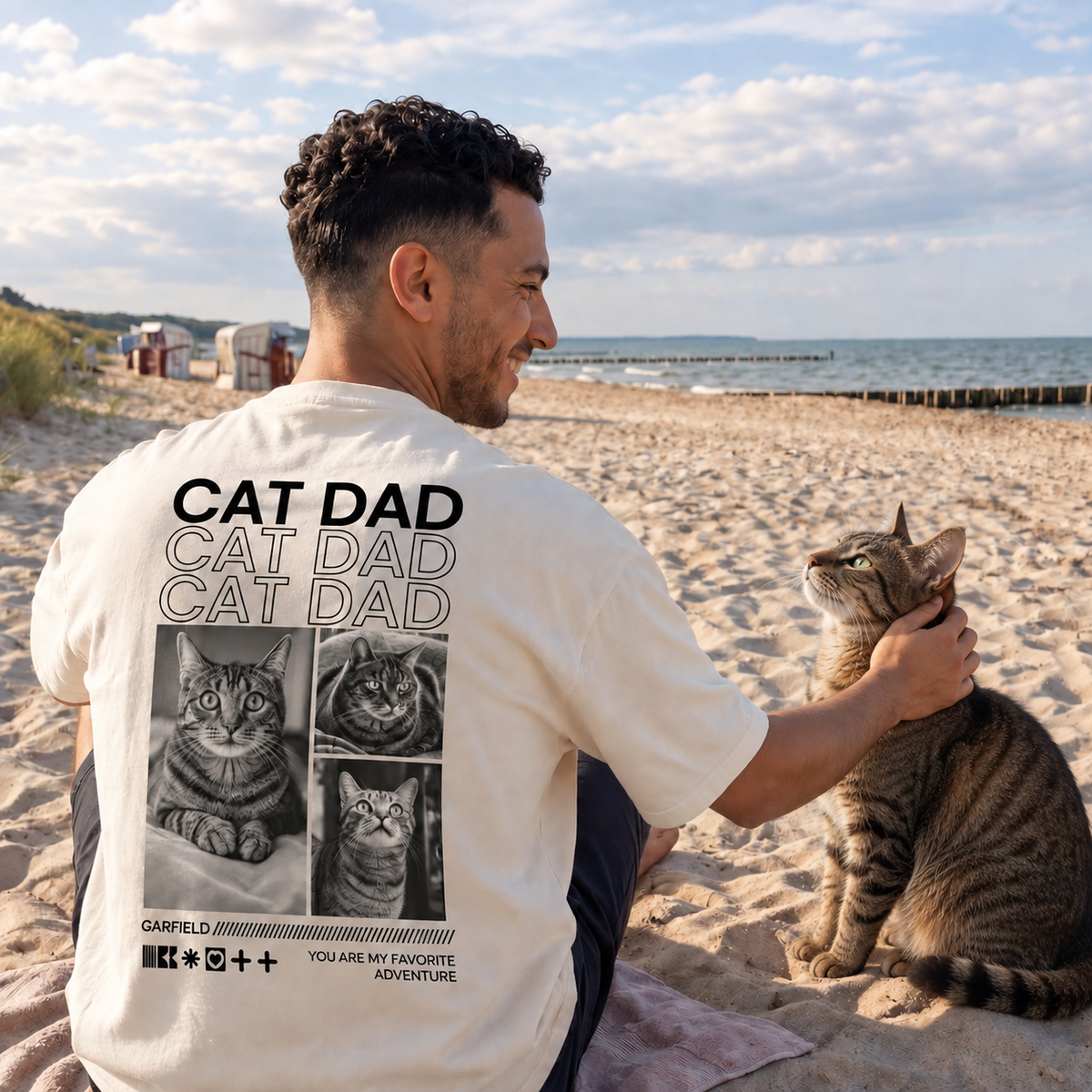 T-shirt beige con la scritta "Cat Dad" e motivi di gatti sul retro, indossata da un uomo in spiaggia.