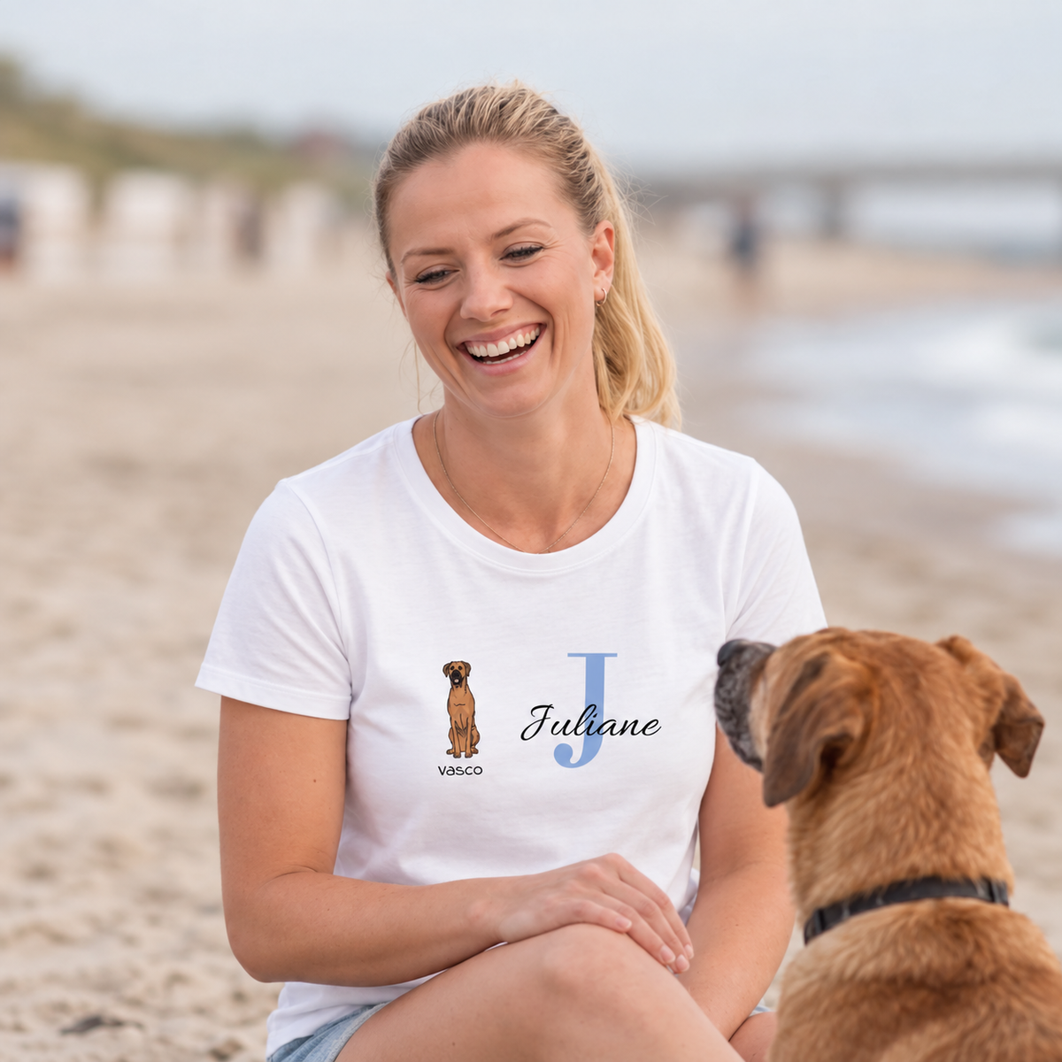 T-shirt bianca con grafica di cane e il nome Juliane in lettere blu. Donna seduta in spiaggia.