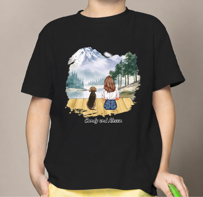 T-shirt per bambini nera con grafica di una bambina e un cane al lago con montagne sullo sfondo.