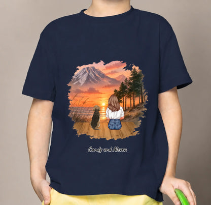 T-shirt per bambini blu con grafica di una persona seduta e un cane in un paesaggio al tramonto.