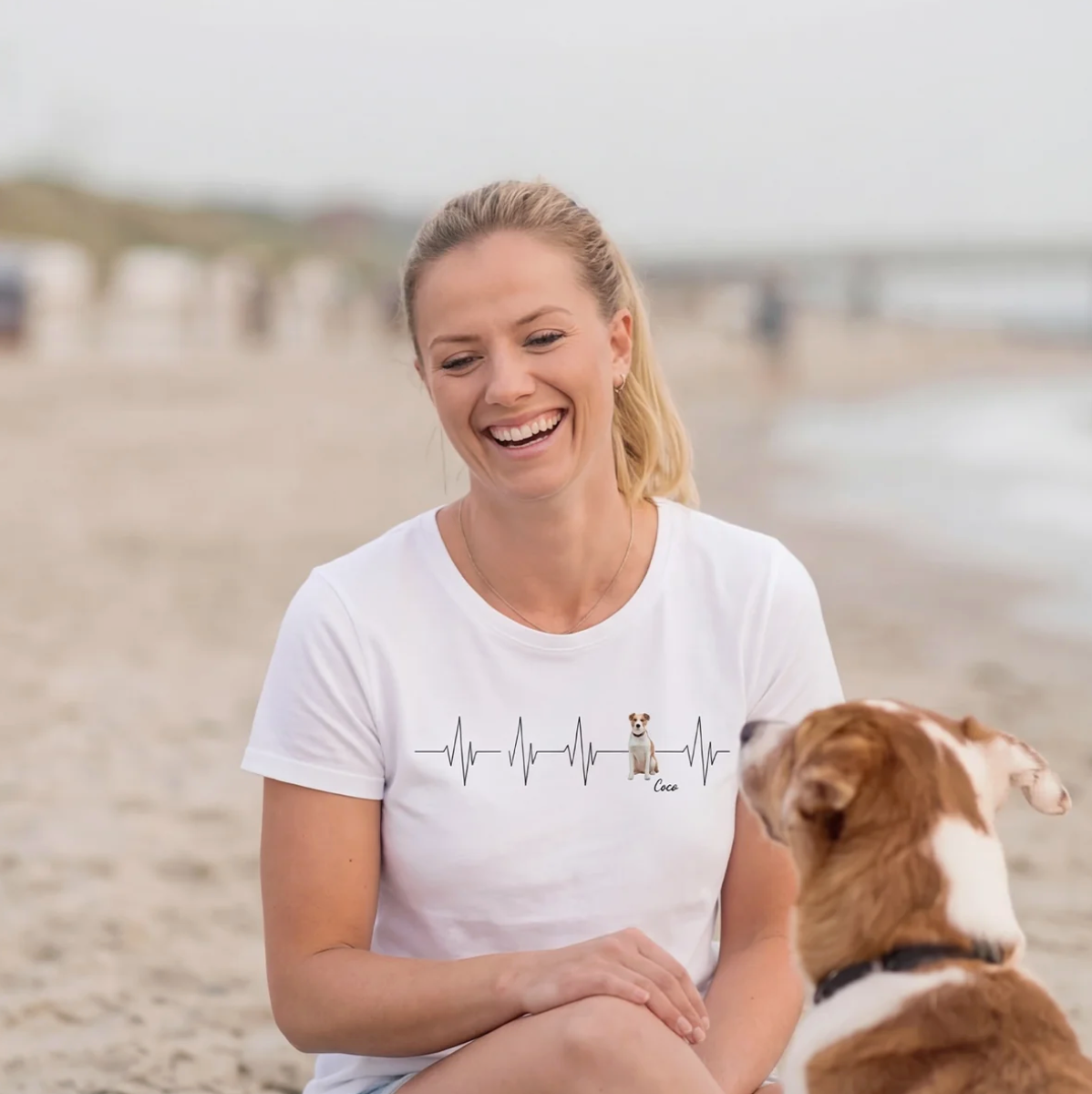 T-shirt bianca con motivo battito cardiaco e cane, donna seduta in spiaggia.
