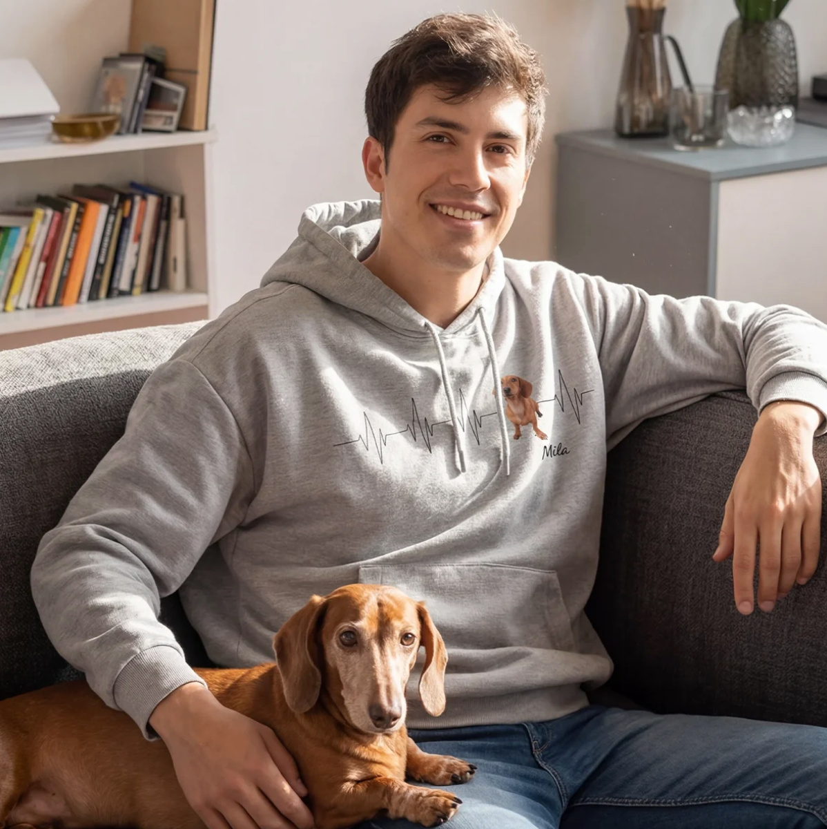 Felpa con cappuccio grigia con motivo battito cardiaco e foto di cane, seduto su un divano con un bassotto in braccio.