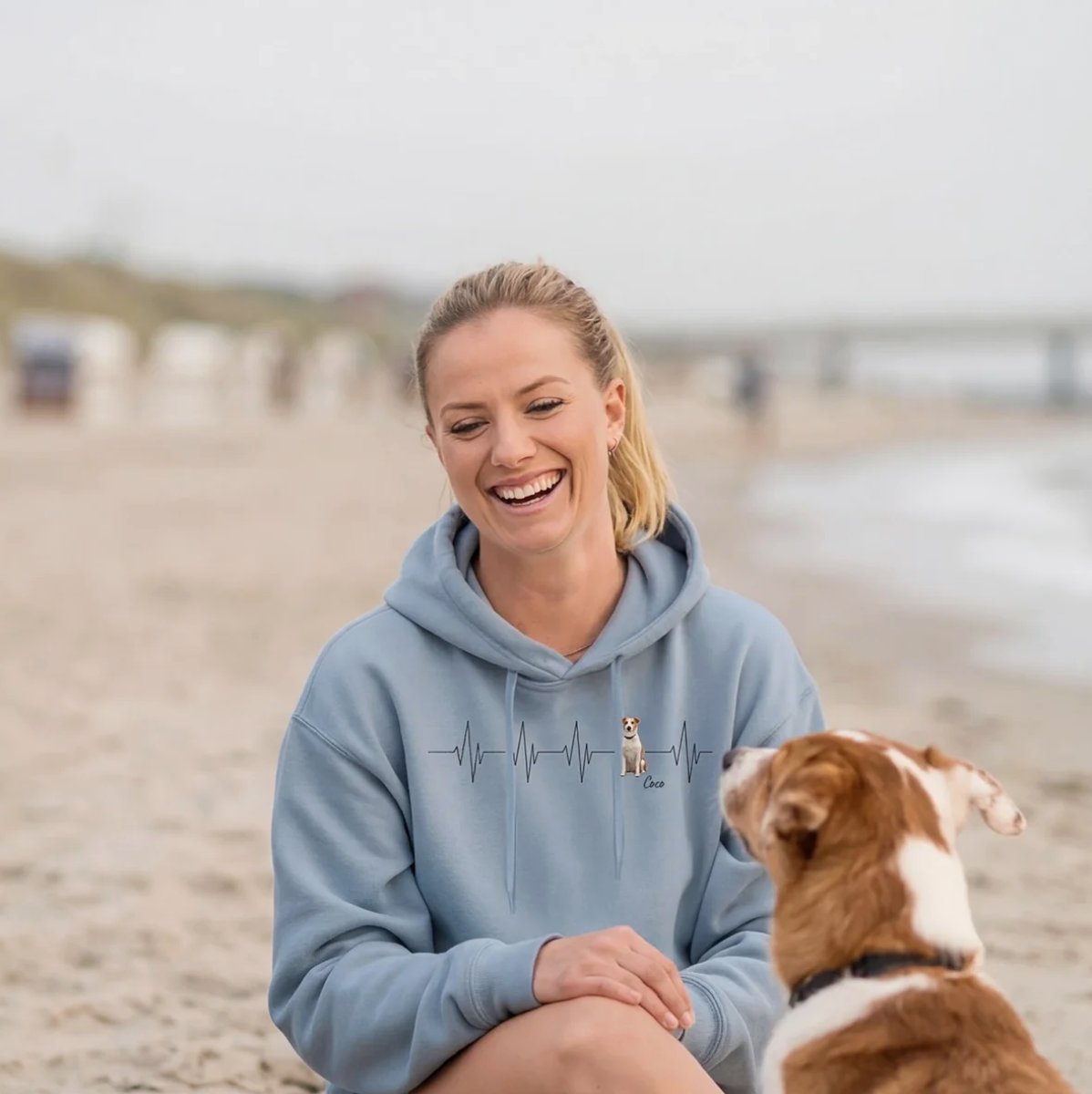 Felpa con cappuccio blu personalizzata con motivo battito cardiaco e cane, donna sorridente seduta in spiaggia.