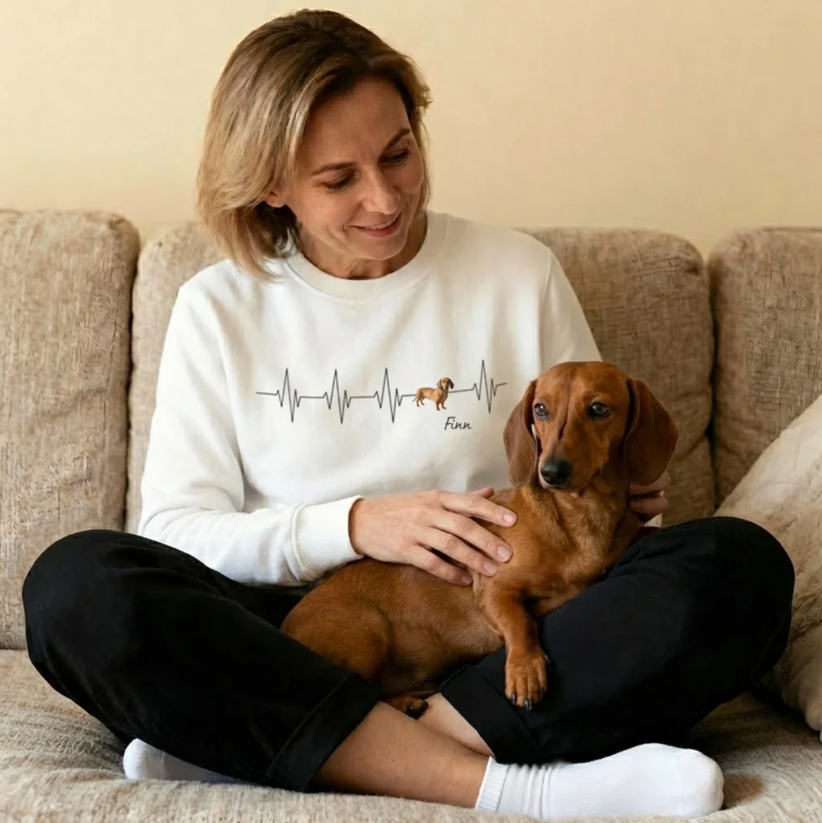 Felpa bianca con motivo battito cardiaco e un cane, donna sorridente seduta su un divano con un bassotto.