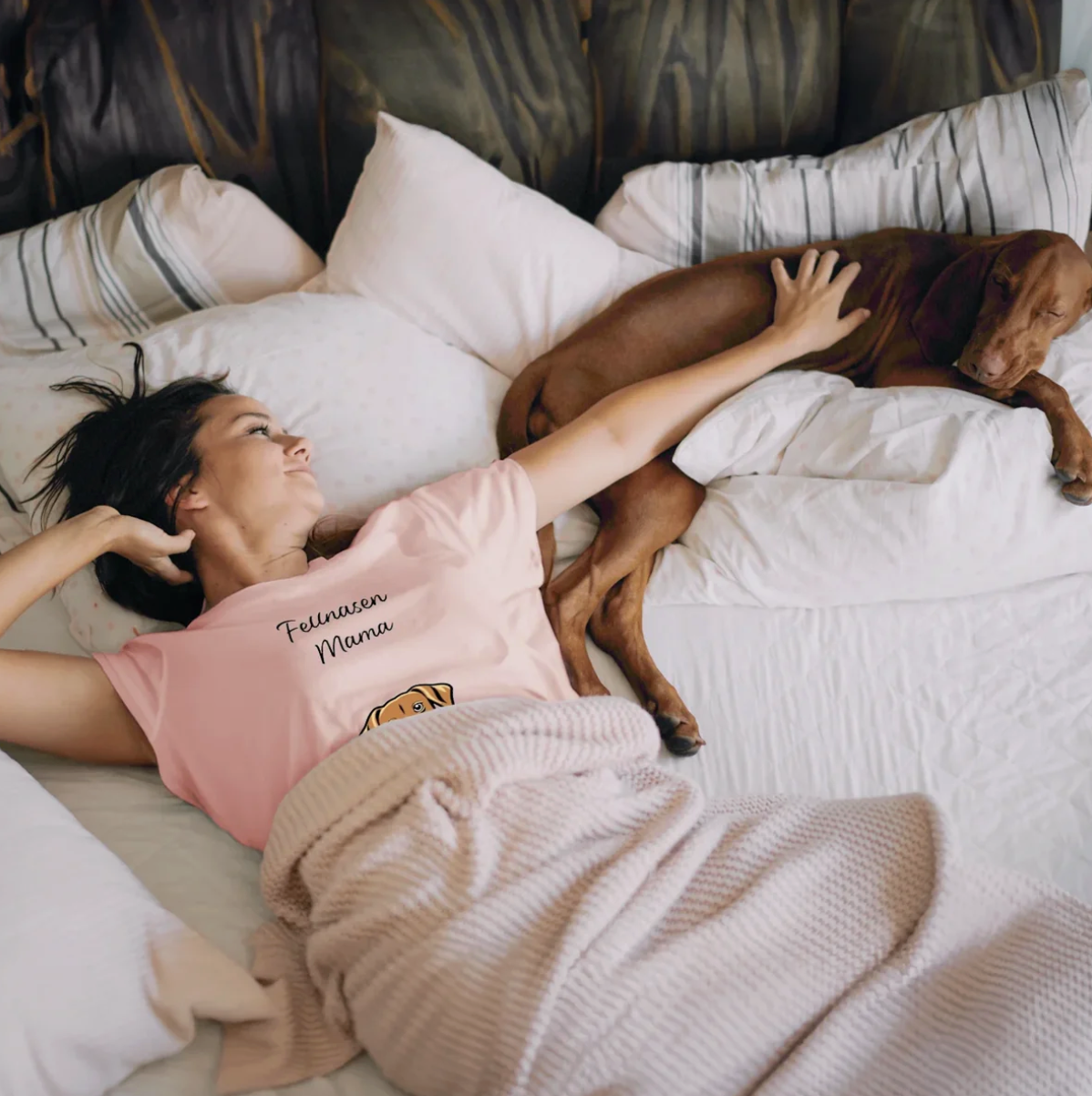 Donna con T-shirt rosa sdraiata sul letto accanto a un cane, circondata da cuscini e una coperta.
