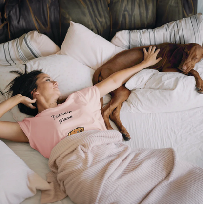 Donna con T-shirt rosa sdraiata sul letto accanto a un cane, circondata da cuscini e una coperta.