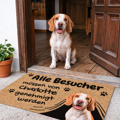Zerbino beige con stampa nera, testo e motivo di cane, davanti a una porta d'ingresso. Cane seduto accanto.