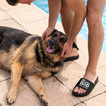 Persona che indossa ciabatte nere con la stampa "THE WALKING DAD", seduta accanto a un cane a bordo piscina.